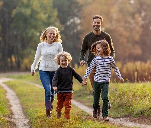 Zusammenfassung: Eine glückliche Familie mit zwei kleinen Söhnen rennt lachend einen Feldweg entlang, umgeben von herbstlicher Natur.  Detaillierte Beschreibung:      Personen: Das Bild zeigt eine Familie, bestehend aus Vater, Mutter und zwei Söhnen. Alle Familienmitglieder wirken fröhlich und lachen. Die Mutter hat blonde, gewellte Haare und trägt einen weißen Zopfstrickpullover, blaue Jeans und braune Stiefel. Der Vater hat dunkles Haar und trägt einen grünen Strickpullover und braune Hosen. Die Söhne, etwa im Vorschulalter, halten sich an den Händen und rennen vor ihren Eltern her. Einer der Jungen hat lockiges blondes Haar und ist in einen grauen Pullover und rote Hosen gekleidet, der andere Junge hat längere braune Haare und trägt ein blau-weiß gestreiftes Oberteil und grüne Hosen.     Umgebung: Die Familie befindet sich inmitten einer herbstlichen Landschaft. Ein schmaler Feldweg schlängelt sich durch das Bild. Beidseitig des Weges erstrecken sich Wiesen mit vereinzelten Gräsern und Pflanzen. Im Hintergrund sind Bäume mit buntem Herbstlaub zu erkennen. Die Farben sind warm und reichen von Gold über Orange bis hin zu Braun.     Atmosphäre und Stimmung: Die Atmosphäre ist fröhlich, unbeschwert und harmonisch. Das Lachen der Familie und die Bewegung im Bild vermitteln ein Gefühl von Freude und Verbundenheit. Das warme Licht und die herbstlichen Farben tragen zu einer positiven und behaglichen Stimmung bei. Das Bild strahlt eine gewisse Natürlichkeit und Authentizität aus und vermittelt den Eindruck eines glücklichen Familienmoments.