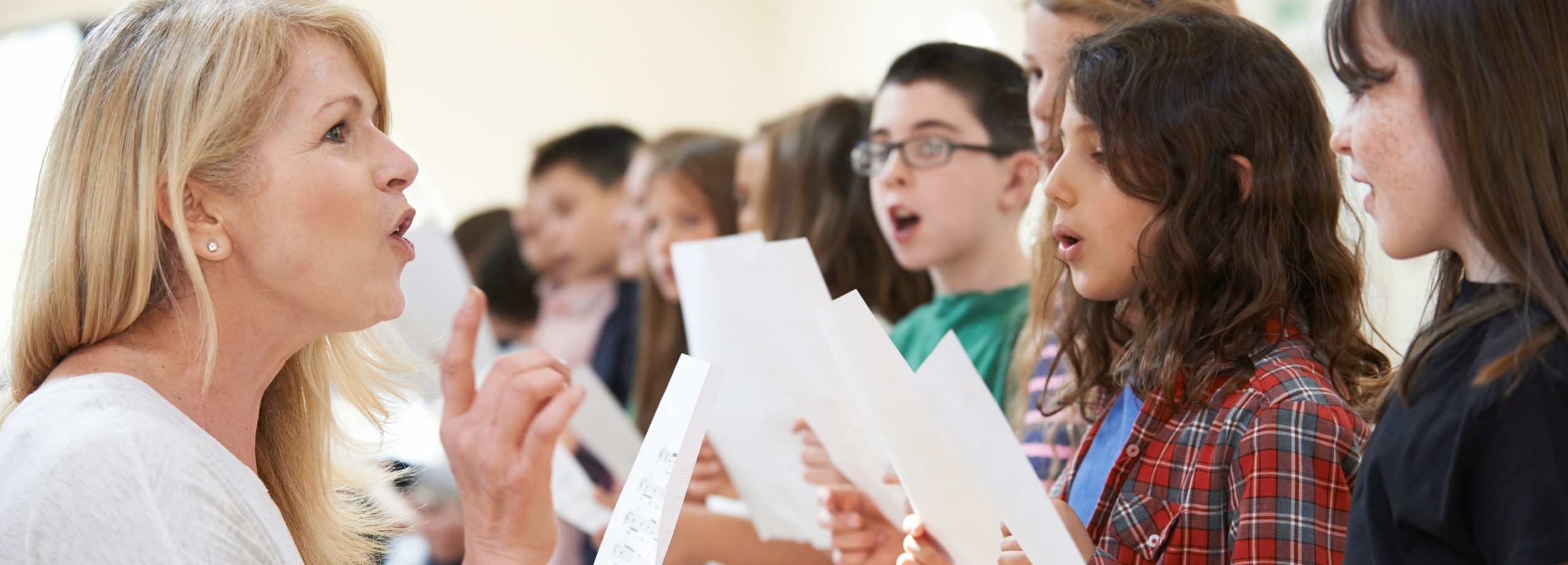 Eine Frau mit blonden Haaren, die ein weißes Oberteil trägt, steht links im Bild und hält eine Hand mit erhobenem Zeigefinger. Sie spricht zu einer Gruppe von Personen, die ihr gegenüberstehen und jeweils ein Blatt Papier in den Händen halten. Die Personen sind in einer Reihe aufgestellt und blicken zur Frau. Eine Person trägt ein rot-kariertes Hemd. Die Frau gibt Gesangsunterricht.